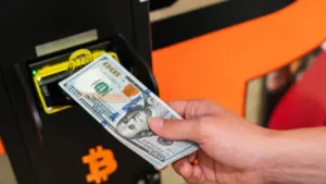 Person inserting a $100 bill into a Bitcoin ATM to buy cryptocurrency, with a Bitcoin logo in the background.